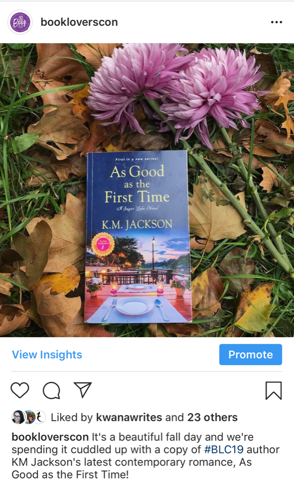 A screenshot features a photo of KM Jackson's As Good as the First Time laying on the grass alongside two purple flowers that is posted on Book Lovers Con's Instagram page.