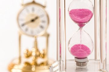 an hourglass filled with pink sand sits in the foreground while a brass clock sits in the background